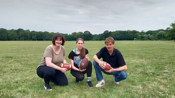 Die Volontäre Corinna, Valerie und Tim sitzen im Park mit einem Football in der Hand.