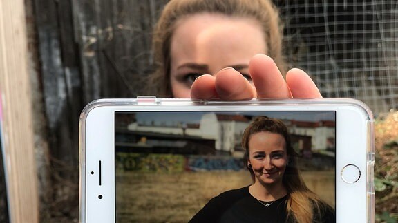 Die von der Sonnen beschienene Johanna Streicher steht vor einer Industrielandschaft und hält ihr Handy in die Kamera