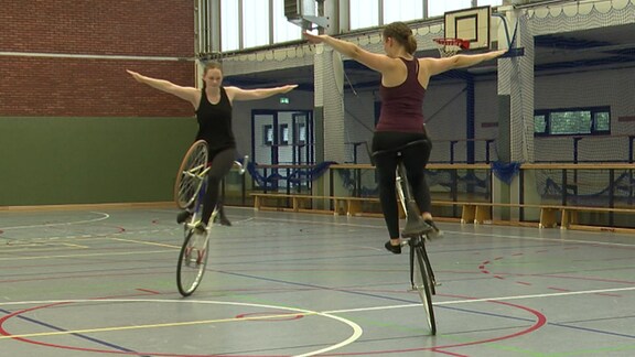 Zwei Frauen fahren in einer Turnhalle Fahrrad im kreis. Sie fahren beide auf den Hinterrädern und sietzen auf dem Lenker.