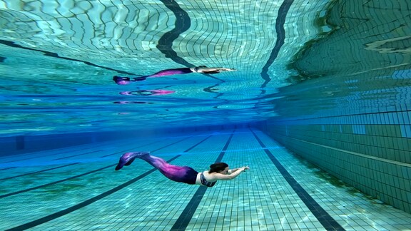 Eine junge Frau schwimmt als Meerjungfrau unter Wasser in einem Schwimmbad. Die Flosse ist violett, auf dem Boden sieht man die Bahnen des Beckens.