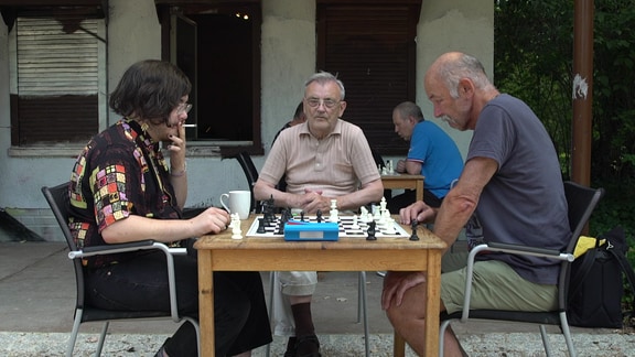 Zwei Männer sitzen sich gegenüber und spielen Schach. Der Linke hält eine Zigarette in der Hand. Zwischen ihnen steht ein Schachbrett. Dahinter sieht man einen weiteren Mann, der ihnen beim Spielen zusieht.