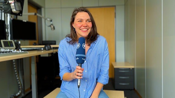 Die Volontärin Henrike Freytag sitzt in einem Radiostudio und hält ein blaues ARD Mikrofon in der Hand.
