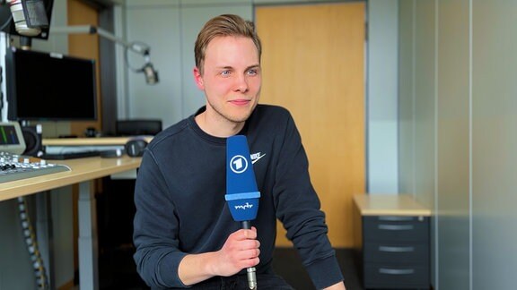 Volontär Tim Magas sitzt mit einem blauen ARD-Mikrofon in einem Radiostudio.