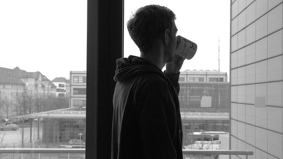 Ein junger Mann steht wartend vor dem Fenster und trinkt aus einer Tasse.