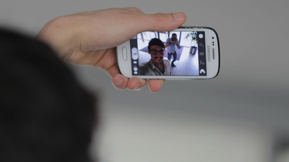 Ein junger Mann fotografiert sich selber mit seinem Handy. Dabei sieht man im Hintergrund einen Fotografen.