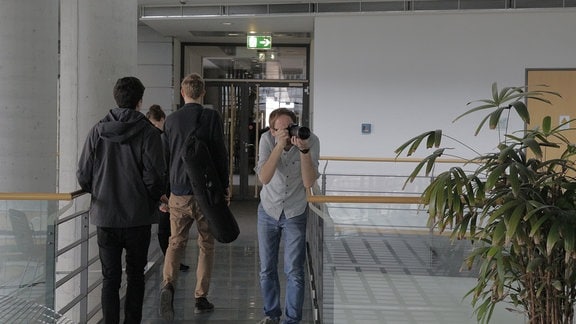 Ein junger Mann fotographiert in die Richtung des Betrachterss. Drei Personen gehen aus der Szenerie, sie tragen Fotoequipment.