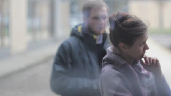 Ein junger Mann und eine junge Frau sind durch ein milchiges Fenster zu sehen. SIe stehen in Jacken draußen vor dem Gebäude. 