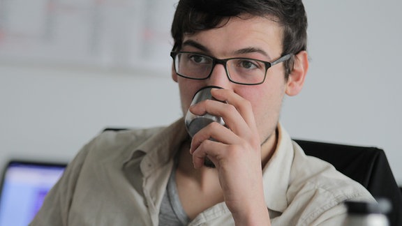 Ein junger Mann mit Brille trinkt aus einem kleinen Metallbecher. 