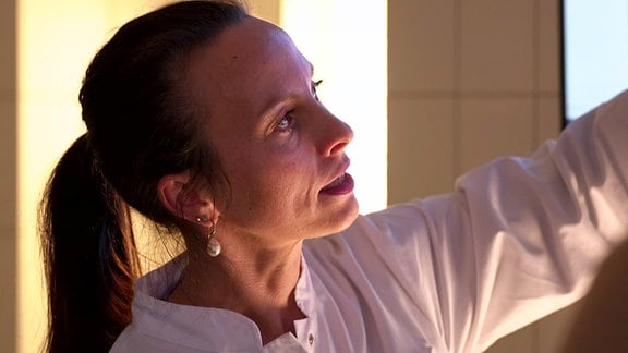 Eine Frau mit langen, zum Zopf gebundenen brünetten Haaren in einem weißen Laborkittel blickt nach oben und hat den linken Arm angehoben, um auf etwas zu zeigen.