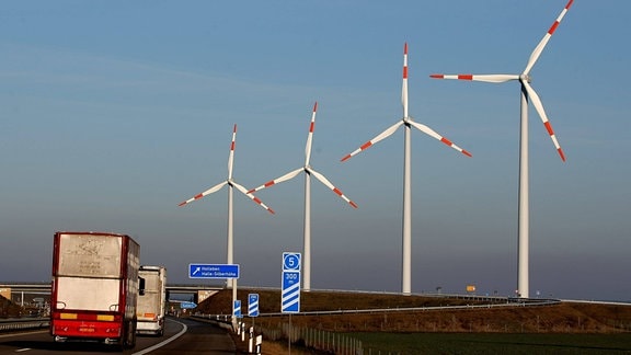 Autobahn 143 mit LKW, Ausfahrts-Meter-Anzeigern und Ausfahrts-Schild nach Holleben und Halle-Silberhöhe, im Hintergrund vier hohe Windräder, Illusion durch Entfernung: Anstieg der Höhe von links nach rechts. Blauer Himmel.