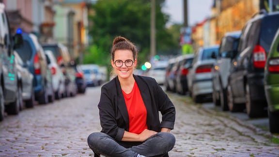 Eine junge Frau sitzt auf der Straße, rechts und links neben ihr parken Autos