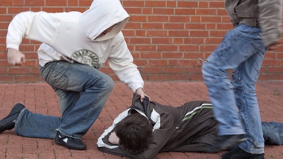 Schlägerei auf dem Schulhof. Ein am Boden liegender wehrloser Junge wird von zwei anderen brutal festgehalten und getreten (gestellte Szene)