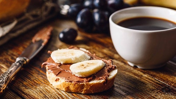 Nahaufnahme Frühstückstisch: Brotscheibe mit Nugataufstrich und Bananenscheiben, Messer mit Nugatrest, daneben gefüllte Kaffeetasse, unscharf im Hintergrund Trauben