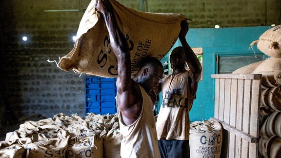 Ein Arbeiter hilft in einem Raum einem anderen Arbeiter, einen schweren Sack mit Kakaobohnen auf den Rücken zu heben, im Hintergrund viele weitere Säcke.