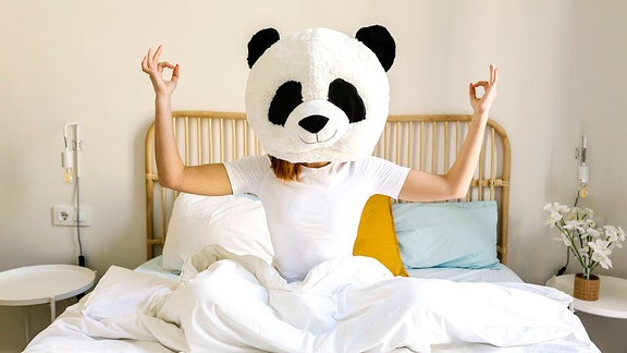 Frau mit Panda-Maske meditiert auf dem Bett zu Hause.