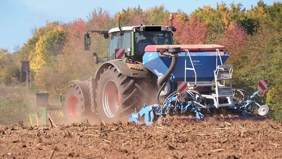 Trockener Acker mit großem Traktor samt angehängtes Gerät. Im Hintergrund ein paar gefärbte Laubbäume- Aufnahme aus Bodennähe.