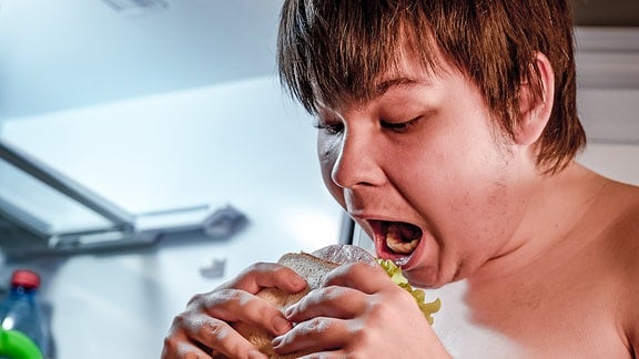 Ein dicker Junge steht vor einem offenen Kühlschrank und beißt in ein Sandwich