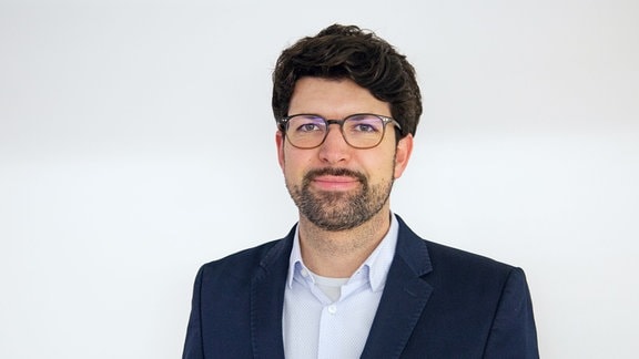 Porträtfoto von Professor Adrian Meier, der ein Mann mit dunklen Locken ist, und Brille und Anzug trägt.