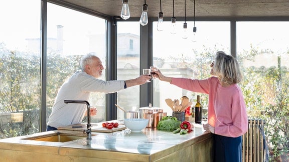 Älteres Paar stößt über Küchentheke hinweg miteinander an.