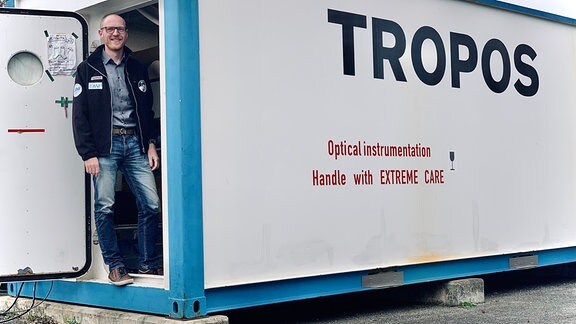 Mann mit Brille und Jacke mit Logos von Tropos und AWI sowie Überwinterungs-Missions-Logo steht in Tür eines weißen Containters mit Aufrischt Tropos und rundem Fenster in Tür