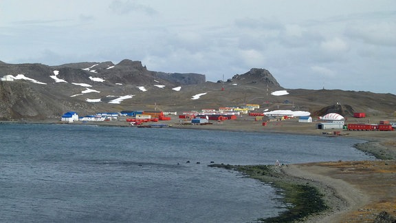 Blick auf den einem Container-Dorf ähnelnden Ort Villa Las Estrellas an einer Bucht auf der König-Georg-Insel in der Antarktis. Grau-brauner Boden, vereinzelt Schnee.