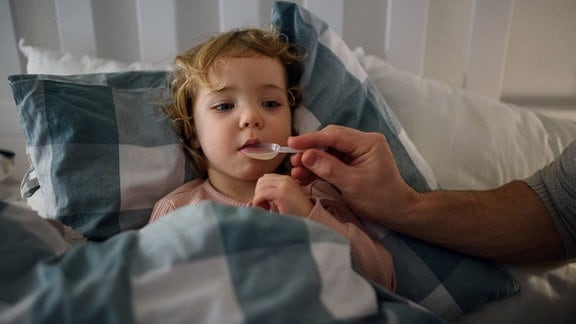 Im Bett liegendes Kleinkind erhält flüssige Medizin auf einem Löffel.