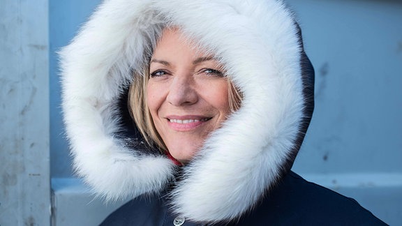 Frau in dicker Winterjacke mit fellbesetzter Kapuze lächelt in die Kamera 