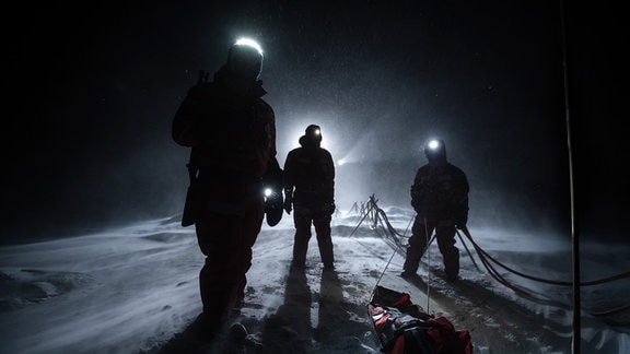 Drei Personen mit Stirnlampen im Dunkeln. Wegen des Gegenlichts aus einem Strahler sind nur ihre Silhouetten zu erkennen. Sie stehen auf einem schneebedeckten Boden neben einem "Handlauf" aus drei Leitungen, die den Weg markieren.