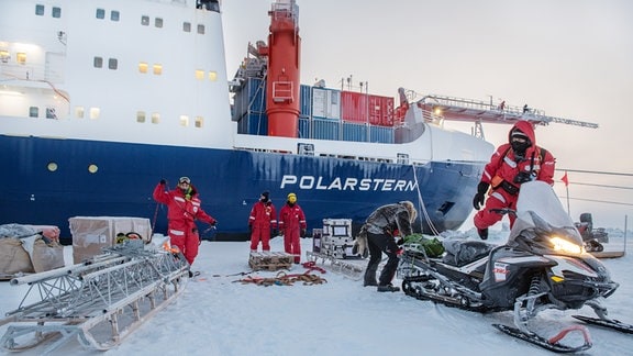 Das Schiff Polarstern wird bei Tageslicht in der Arktis entladen. Im Hintergrund das im Eis festsitzende Schiff. Davor fünf Forscher*innen, die Equipment enladen haben und auf zwei Schlitten platzieren, die von Schneemobilen gezogen werden.