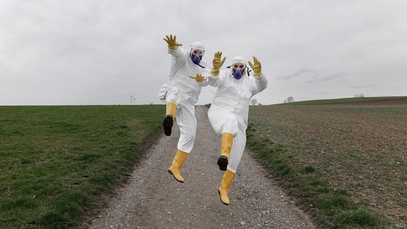 Zwei Personen mit Schutzanzug, Schutzbrille, Maske und Gummistiefeln springen zueinander auf einem Feldweg, Frontalansicht