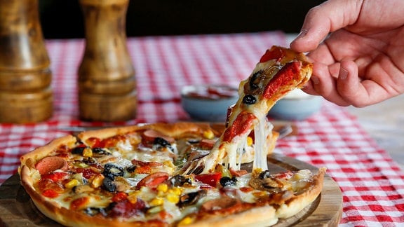 Ein fettiges Stück Pizza wird von einer Hand aus der Pizza gehoben und zieht Käse-Fäden, rot-weiß-karierte Tischdecke, Salz- und Pfeffer-Streuer.