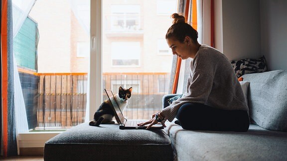 Frau (Freiberuflerin) sitzt in gemütlicher Kleidung auf Sofa mit Laptop, dahinter Katze. Seitliche Aufnahme, im Hintergrund Fenster und Balkon.