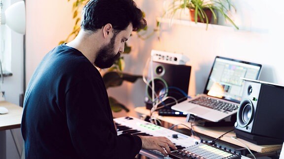 Mann sitzt im heimischen Musikstudio an Tasteninstrument und Mischpult, mit Laptop und Boxen. Aufnahme von schräg hinten, kontrastreiche Farben, Tiefenunschärfe.