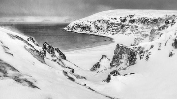 Schwarz-weiß-Bild von felsiger, schneebedeckter, karger Landschaft an Meer; leichtes Schneetreiben