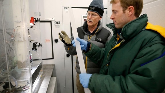 Zwei Wissenschaftler mit warmen Jacken begutachten in einem Labor einen langen Stab aus Eis.