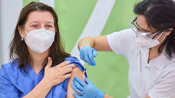 Eine Frau in einem blauen Krankenhauskittel erhält eine Impfjng von einer zweiten Frau in weißer Kleidung. Beide Frauen tragen FFP-2-Masken.