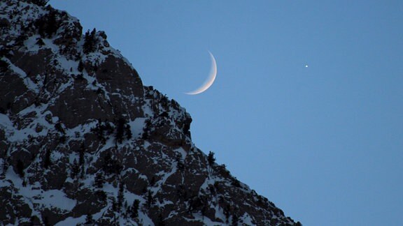 Die Mondsichel hinter einem Berg. Neben ihr einer der Planeten in unserem Sonnensystem.