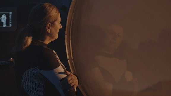 Raumfahrerin schaut mit verschränkten Armen leicht melancholigshc aus Fenster einer Mars-Basis nach draußen. Ansicht seitlich von hinten.