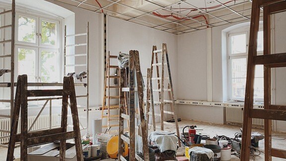 In einem hellen Raum mit zwei über Eck liegenden Fenstern stehen viele Leitern, Eimern, Gerüstteile und sonstige Baumaterialien