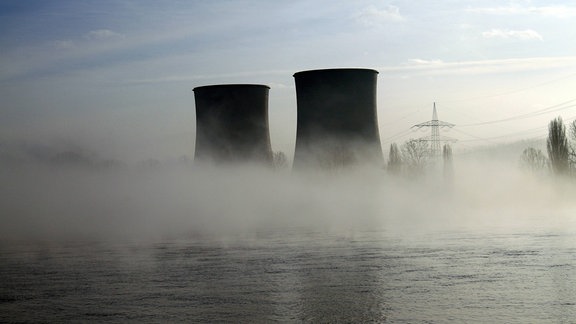 Zwei dunkle Kühltürme eines Atomkraftwerks schauen aus bodennahen Nebel heraus. Im Vordergrund Wasser, im Hintergrund Hochspannungsleitung, Bäume und heiterer Himmel.