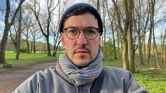 Mann mit Schal, Mütze und Brille blickt in die Kamera, steht dabei auf einem Weg in einem Park
