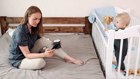 Junge Mutter sitzt mit einer Kaffeetasse auf dem Bett und beantwortet Nachrichten am Telefon, während das Baby im Kinderbett steht.