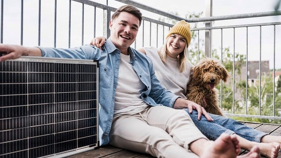 Fröhliches Paar aus jungem Mann und junger Frau sitzt mit Hund auf Balkon neben einem Solar-Paneel, auf das Mann seinen Arm gelegt hat, Balkon-Brüstung aus Metall, Blick auf die Stadt dahinter