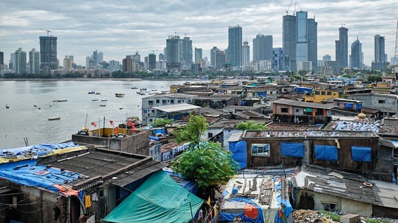 Mumbai: Slum-ähnliche Siedlung im Vordergrund, Skyline im Hintergrund, an Wasser gelegen, bewölkter Himmel