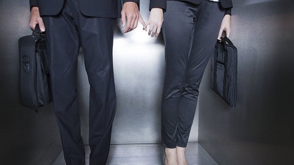 Eine Frau und ein Mann (nur Oberkörper und Beine zu sehen) in formeller Berufskleidung stehen in einem Aufzug und berühren sich unauffällig mit den Fingern.