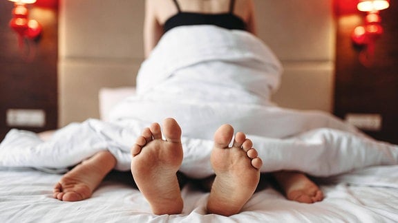 Zwei Personen übereinander in einem Bett, Ansicht vom Fußende aus, bedeckt mit Decke, unscharf im Hintergrund Oberkörper einer Frau mit Top.