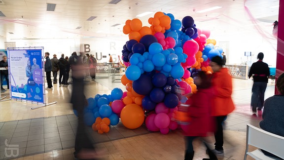 In einem alten Kaufhaus steht eine große Skulptur aus Ballons. Daneben sind unscharf Menschen zu sehen.