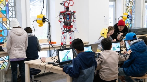 Mehrere Kinder sitzen an Laptops. In ihrer Mitte steht Roboter in Form eines Menschen.