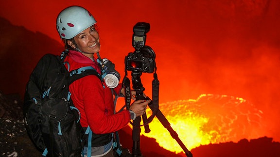 Eine Frau mit Helm, Rucksack und einer großen Fotoausrüstung in der Hand steht vor rot leuchtender Lava und schaut in die Kamera.