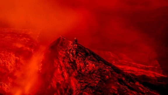 Eine Person sitzt auf einem Felsen und blickt hinunter zu roter Lava.